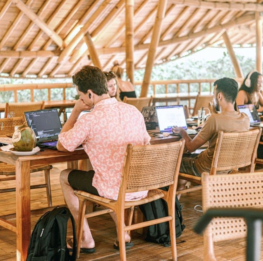 Interns working on laptops at a long shared table in an open bamboo coworking space in Bali