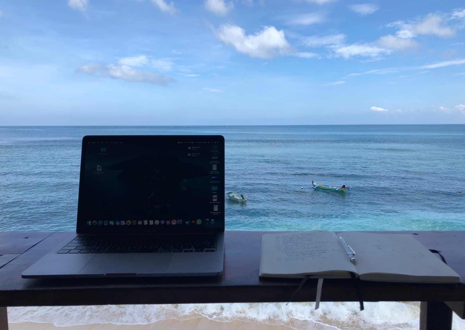 A laptop and an open notebook with a pen resting on a wooden ledge overlooking the sea in Bali, with small fishing boats moored on the water
