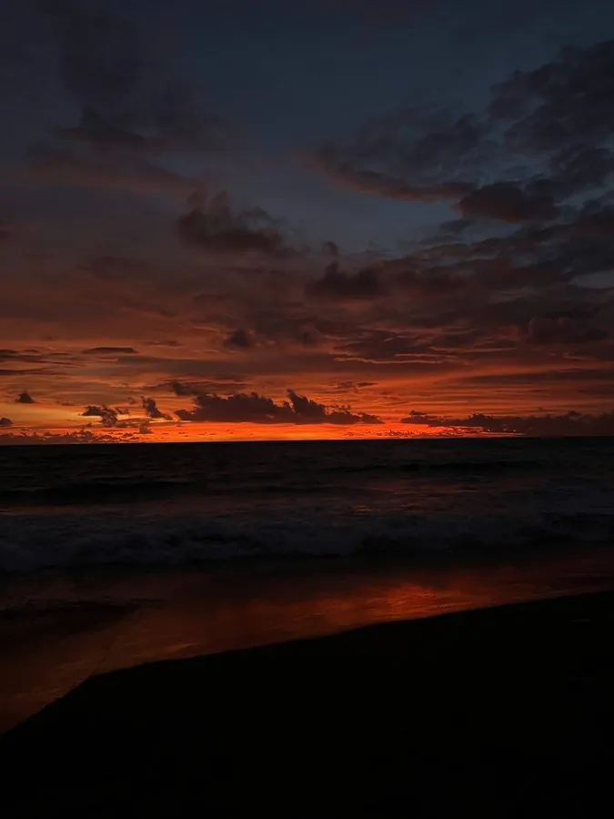 Sunset over the sea from a dark volcanic-sand beach in Bali, with orange light breaking under the cloud