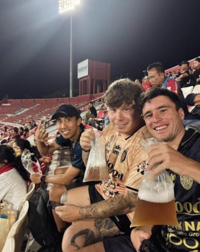 Interns at a Bali United football match at night, holding drinks in the stand