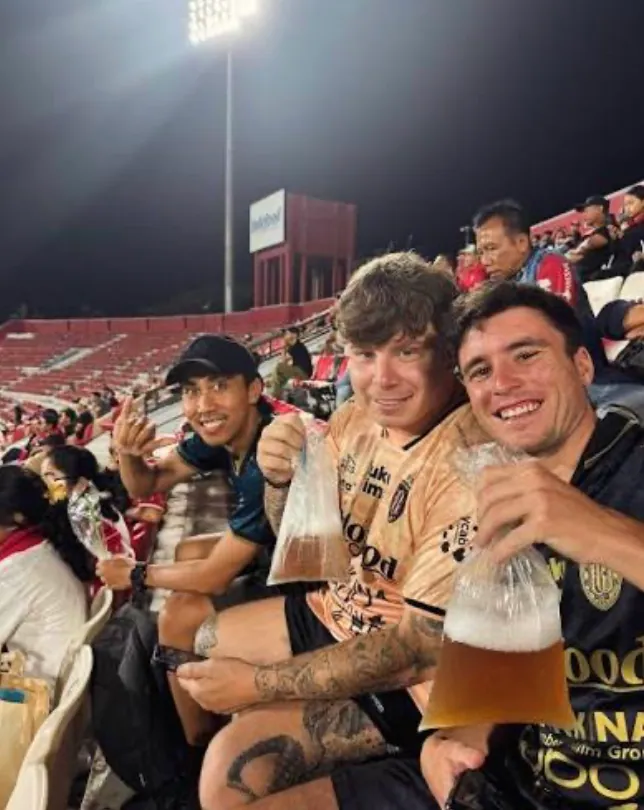 Interns at a Bali United football match at night, holding drinks in the stand