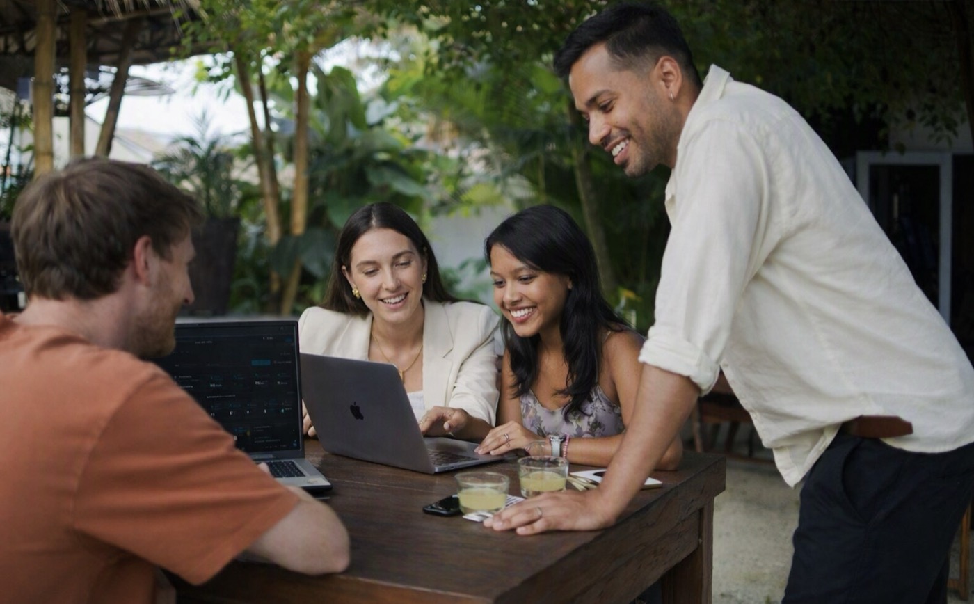 Four interns and a colleague gathered around laptops at an outdoor table in Bali, reviewing work together