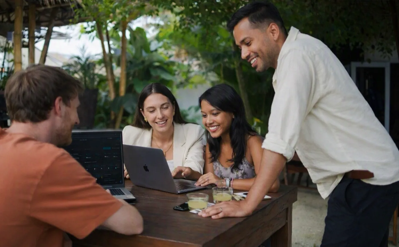 Four interns and a colleague gathered around laptops at an outdoor table in Bali, reviewing work together
